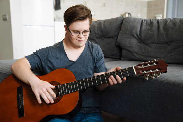 Young man with down syndrome playing guitar while sitting on floor at home - Foto, afbeelding