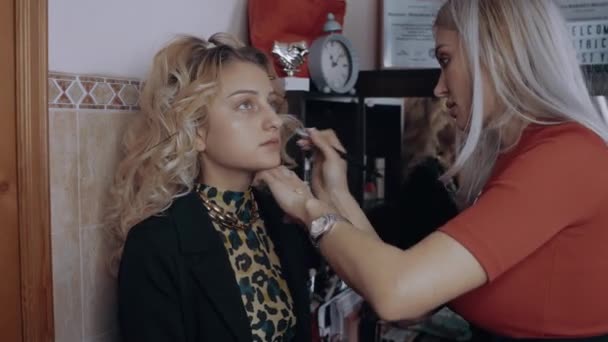 Die blonde Visagistin trägt die Schminkbasis der Frauen mit einem Pinsel auf. Das Auftragen von Make-up zu Hause. Künstlerisches Konzept - Filmmaterial, Video
