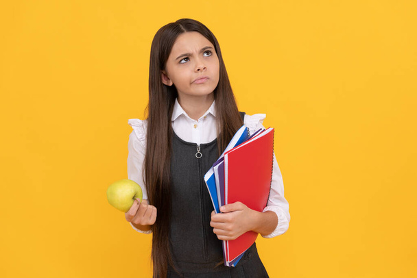 Серьёзные девочки школьного возраста думают, держа яблоко и книги на желтом фоне, думая: - Фото, изображение