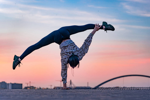 Silhouette of flexible female circus artist doing handstand on the dramatic sunset and cityscape. Concept of individuality, creativity and outstanding - Foto, Imagen