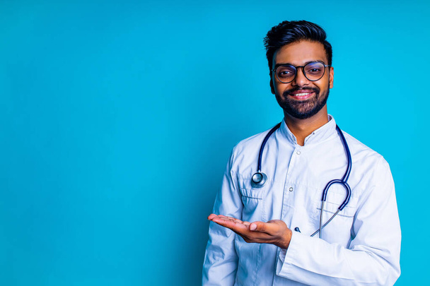 pakistanian doctor in white coat with stethoscope pointing at copyspace with hand mock up over blue studio background - Foto, Bild