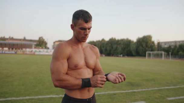 Um homem musculoso faz uma pausa entre exercícios de força no estádio e endireita as pulseiras em seus braços. Movimento lento - Filmagem, Vídeo