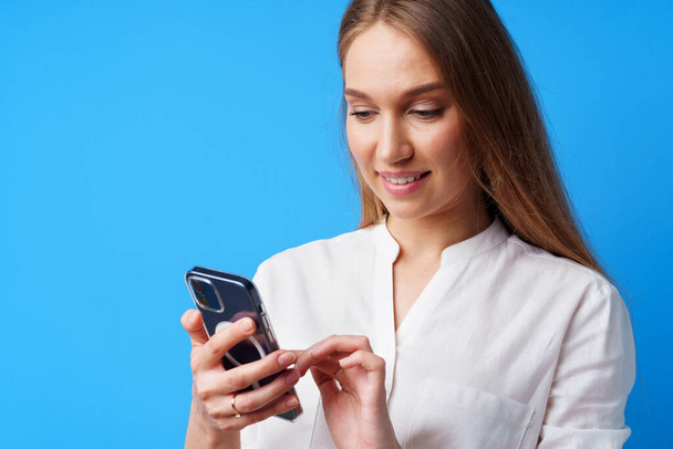 Close up of young blonde woman using her smartphone against blue background - Фото, изображение