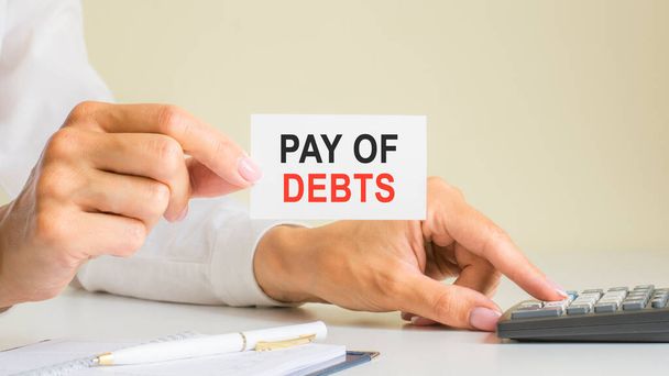 pay of debts, message on business card shown by woman pressing calculator key at workplace in light office, selective focus, business and financial concept - Photo, image