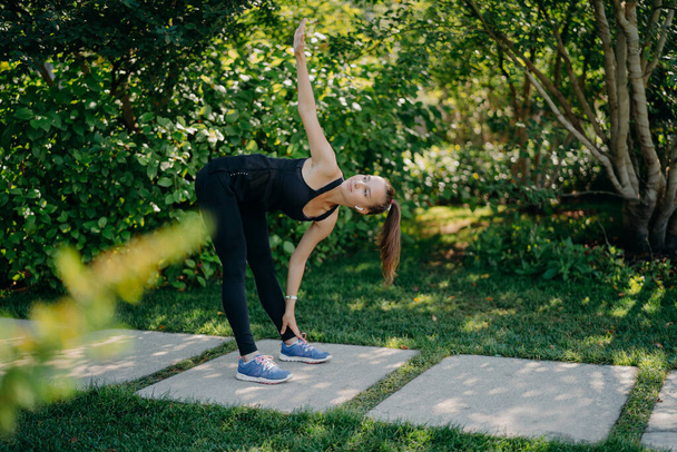 Actieve fitness model oefeningen buiten leunt aan de voeten verhoogt arm bereidt spieren voordat cardiotraining uitdagingen zelf gekleed in actieve dragen heeft ponystaart poses in groen park tijdens de zonnige dag - Foto, afbeelding