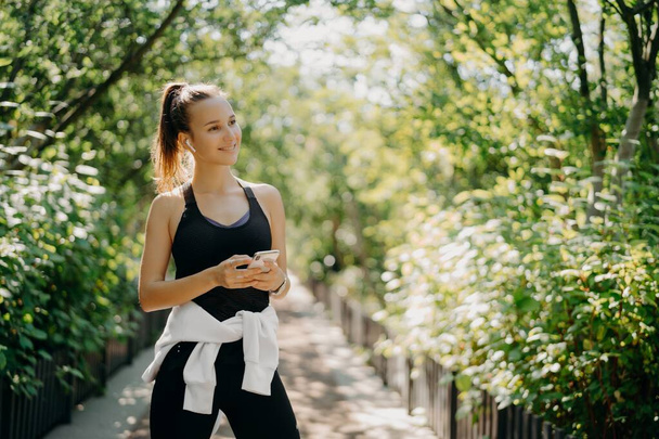 Poziomy strzał aktywnej sportowej kobiety używa smartfona do sprawdzania wyników po jogging ubrany w aktywne zużycie cieszy ciepły słoneczny dzień słucha muzyki przez słuchawki. Koncepcja zdrowego stylu życia. - Zdjęcie, obraz