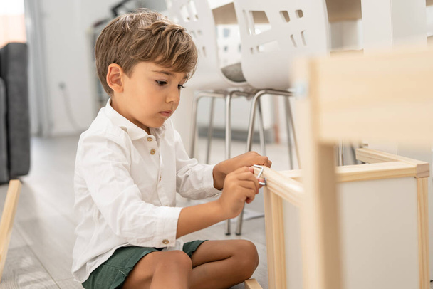 Small caucasian boy four years old playing with self assembly chair or table on the floor at home in day - little child assembling furniture alone childhood craft concept front view copy space - Foto, imagen