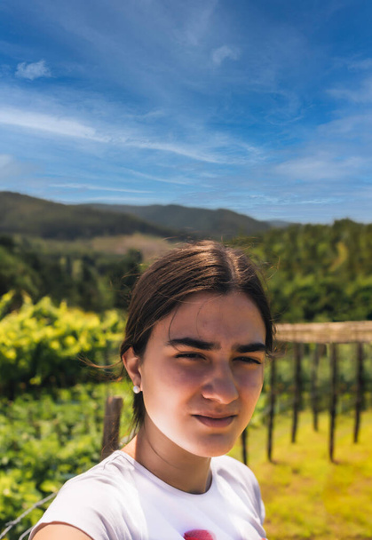 adolescente sosteniendo un teléfono móvil y tomando una selfie con el fondo de un huerto, montañas y un cielo azul en un día de verano con enfoque selectivo. - Foto, imagen