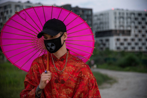 Θετικός άνδρας ντυμένος με ρούχα δρόμου κόκκινο κιμονό με αλυσίδες γύρω από το λαιμό, μαύρο καπέλο και μαύρη ιατρική μάσκα. Νεολαία και τρόπος ζωής. μολυσμένος αέρας πόλης - Φωτογραφία, εικόνα