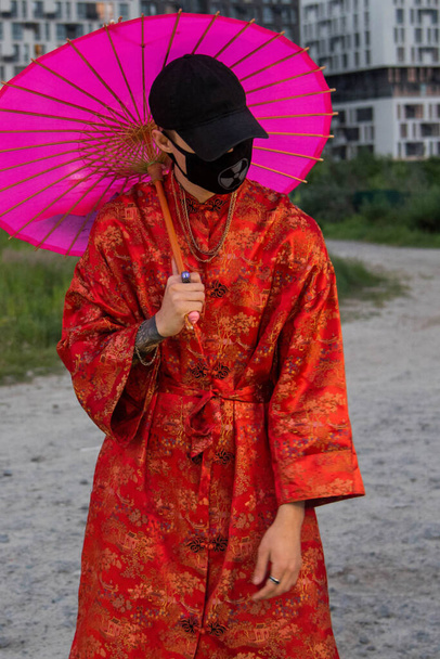 Homme positif habillé dans des vêtements de rue kimono rouge avec des chaînes autour du cou, casquette noire et masque médical noir. Concept de jeunesse et style de vie. air pollué de la ville - Photo, image