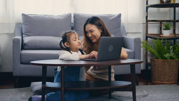 Asian young mother with laptop computer teaching her kid to learn or study online in living room at home, Mum and little preschool daughter learning online on computer, family homeschooling online - Foto, immagini