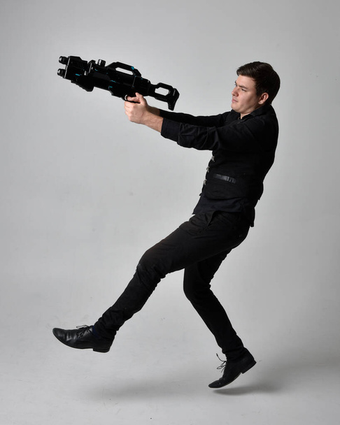 Full length portrait of a  brunette man wearing leather jacket  and holding a science fiction gun.  Standing  action pose isolated  against a grey studio background. - Fotografie, Obrázek