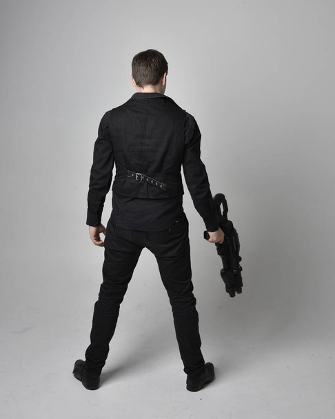 Портрет брюнетки в шкіряній куртці і тримає науково-фантастичний пістолет. Позиція стоячої дії ізольована на сірому фоні студії
. - Фото, зображення