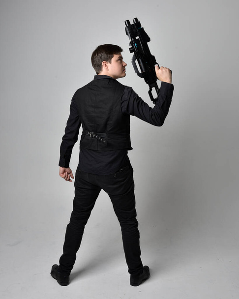 Ganzkörperporträt eines brünetten Mannes in Lederjacke und mit einer Science-Fiction-Waffe. Stehende Action-Pose isoliert vor grauem Studiohintergrund. - Foto, Bild