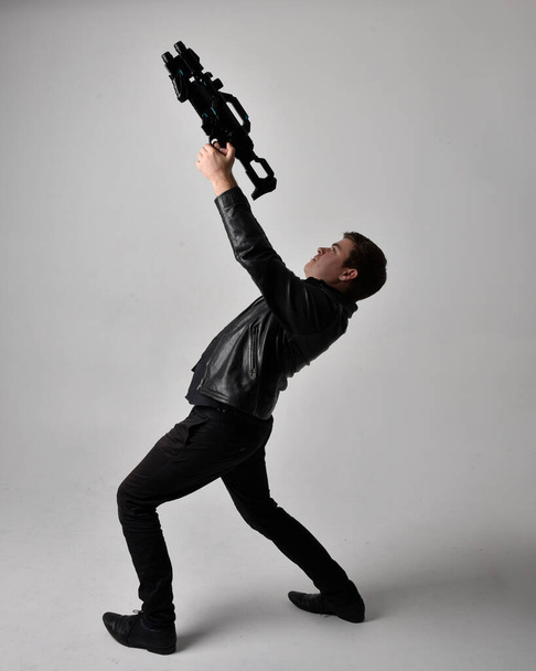Full length portrait of a  brunette man wearing leather jacket  and holding a science fiction gun.  Standing  action pose isolated  against a grey studio background. - Foto, immagini