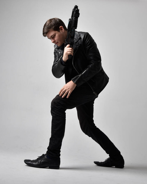 Full length portrait of a  brunette man wearing leather jacket  and holding a science fiction gun.  Standing  action pose isolated  against a grey studio background. - Φωτογραφία, εικόνα