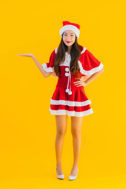 Πορτρέτο όμορφη νεαρή ασιατική γυναίκα χριστουγεννιάτικα ρούχα και καπέλο χαμόγελο ευτυχισμένη με άλλη δράση σε κίτρινο απομονωμένο φόντο - Φωτογραφία, εικόνα