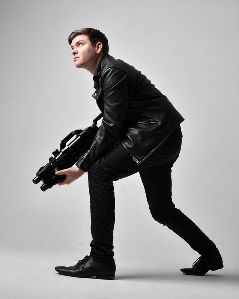 Full length portrait of a  brunette man wearing leather jacket  and holding a science fiction gun.  Standing  action pose isolated  against a grey studio background. - Zdjęcie, obraz