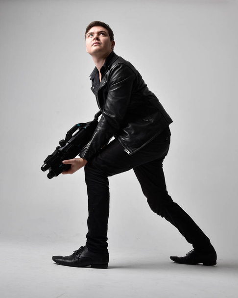 Plný portrét bruneta muže v kožené bundě, který drží sci-fi zbraň. Stojící akční póza izolované na šedém pozadí studia. - Fotografie, Obrázek