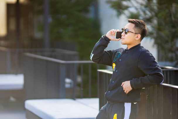 Jonge Aziatische man het dragen van zonneglas en sport jas met behulp van mobiele telefoon na ochtend oefening met kopieerruimte - Foto, afbeelding