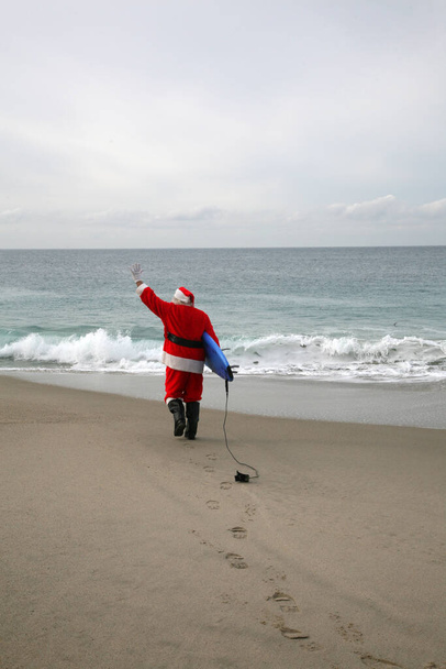 Christmas. Surfing Santa Claus. Surfing Santa. Santa Claus with a surf board and the ocean in the background. Santa Claus loves Surfing when he is not busy delivering presents to boys and girls at Christmas. Santa holding a surfing board. Santa  - Foto, imagen
