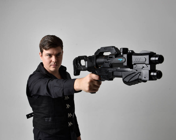 zblízka portrét bruneta muže v kožené bundě a drží sci-fi zbraň. akční póza izolovaná od šedého pozadí studia. - Fotografie, Obrázek