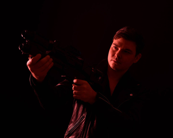 közeli portré egy barna férfiról bőrdzsekiben és sci-fi pisztollyal a kezében. Állandó akció póz piros sziluett világítás ellen egy fekete stúdió háttér. - Fotó, kép