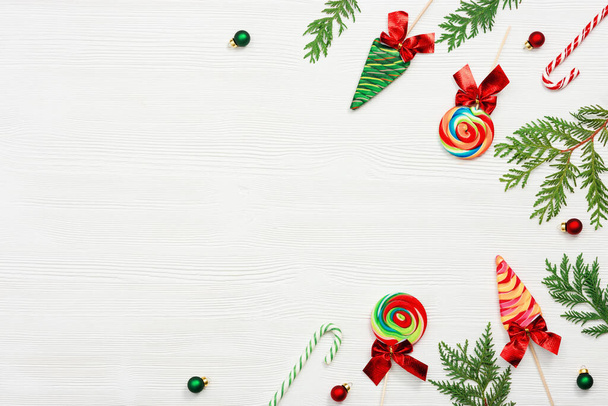 Christmas sweets assorted top view, green thuja branches, small red and green ball toys on white wood background, flat lay. Bright New Year candy and lollipops. - Foto, afbeelding