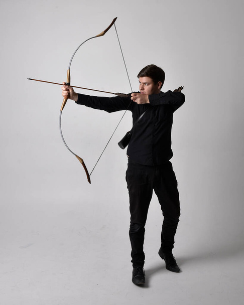 Full length portrait of a  brunette man wearing black shirt and waistcoat holding  a bow and arrow.  Standing  action pose isolated  against a grey studio background. - Foto, imagen