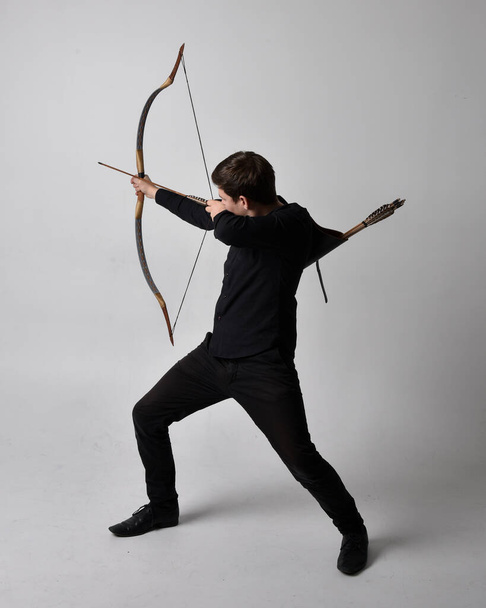 Full length portrait of a  brunette man wearing black shirt and waistcoat holding  a bow and arrow.  Standing  action pose isolated  against a grey studio background. - Zdjęcie, obraz