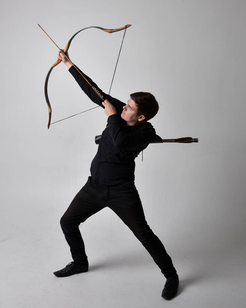 Portrait complet d'un homme brune portant une chemise noire et un gilet tenant un arc et une flèche. L'action debout pose isolée sur un fond de studio gris. - Photo, image