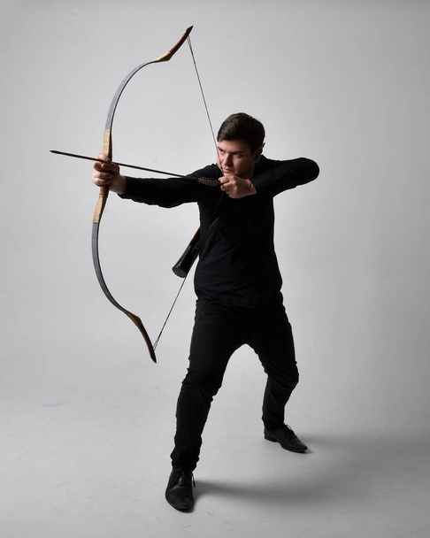 Full length portrait of a  brunette man wearing black shirt and waistcoat holding  a bow and arrow.  Standing  action pose isolated  against a grey studio background. - Photo, Image