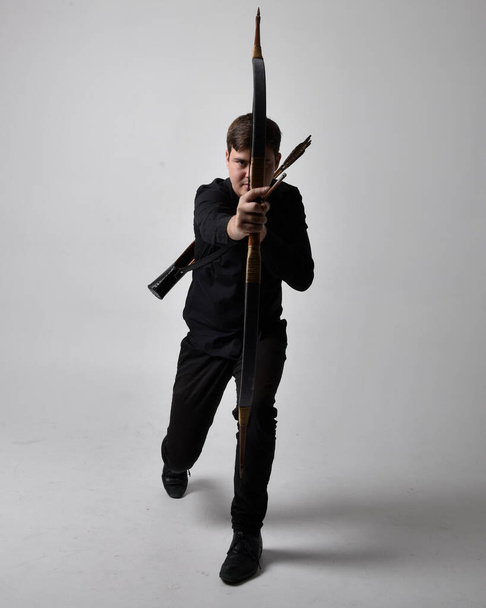 Full length portrait of a  brunette man wearing black shirt and waistcoat holding  a bow and arrow.  Standing  action pose isolated  against a grey studio background. - Photo, Image