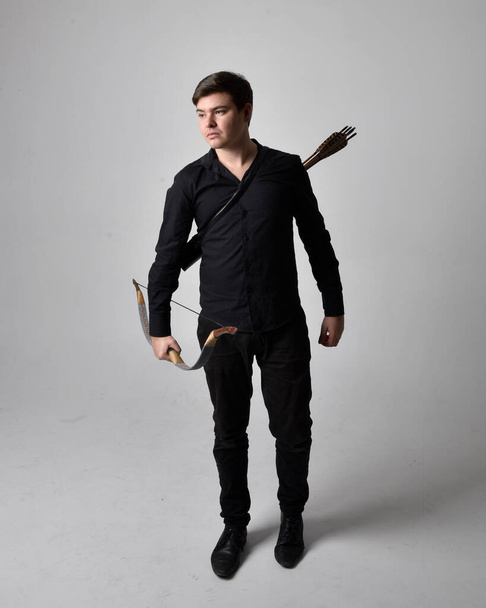 Full length portrait of a  brunette man wearing black shirt and waistcoat holding  a bow and arrow.  Standing  action pose isolated  against a grey studio background. - Foto, Imagem