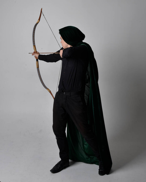 Ολόσωμο πορτραίτο ενός μελαχρινού άνδρα με μαύρο πουκάμισο, γιλέκο και πράσινο βελούδινο μανδύα που κρατάει τόξο και βέλος. Μόνιμη δράση θέτουν απομονωμένη σε ένα γκρίζο φόντο στούντιο. - Φωτογραφία, εικόνα