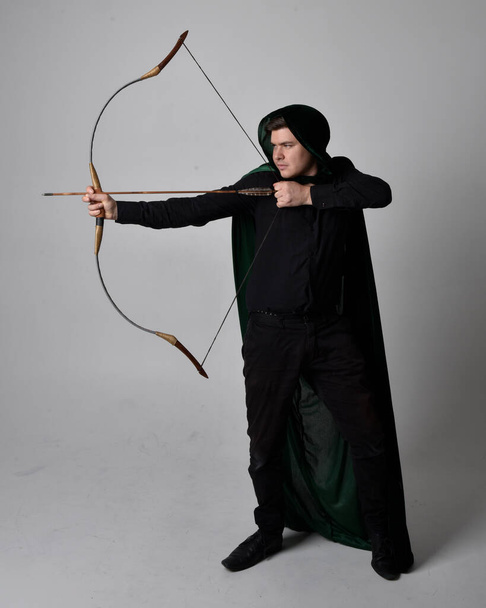 Full length portrait of a  brunette man wearing black shirt, waistcoat and a green velvet cloak holding a bow and arrow.  Standing  action pose isolated  against a grey studio background. - Φωτογραφία, εικόνα