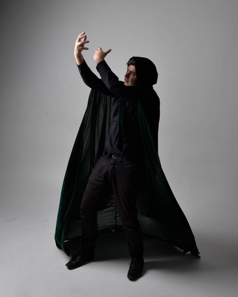 Full length portrait of a  brunette man wearing black shirt, waistcoat and a green velvet cloak.  Standing  action pose isolated  against a grey studio background. - Fotó, kép