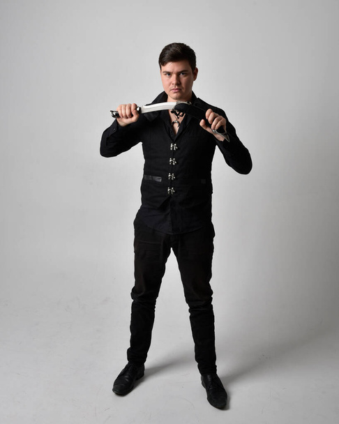 Full length portrait of a  brunette man wearing black shirt and gothic waistcoat holding sword.  Standing  action pose isolated  against a grey studio background. - Valokuva, kuva