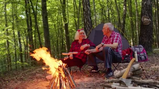Senior elderly grandmother grandfather cooking frying sausages over campfire in wood at camping - Footage, Video