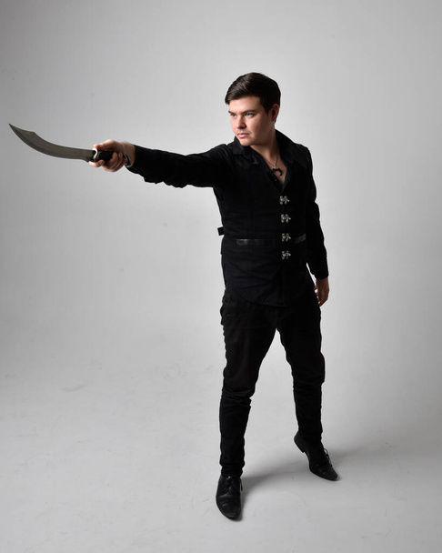 Full length portrait of a  brunette man wearing black shirt and gothic waistcoat holding sword.  Standing  action pose isolated  against a grey studio background. - Foto, immagini