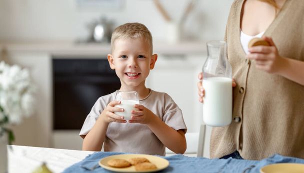 Fröhlicher Junge mit einem Glas frischer Milch lächelt beim Familienfrühstück in die Kamera - Foto, Bild