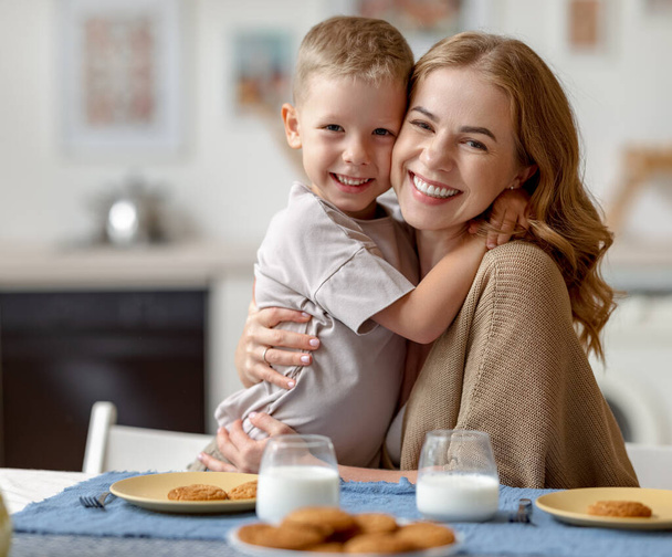 Optimistische vrouw omhelst vrolijke jongen en kijkt naar de camera terwijl zitten aan tafel met koekjes en melk en ontbijten in de ochtend thuis - Foto, afbeelding