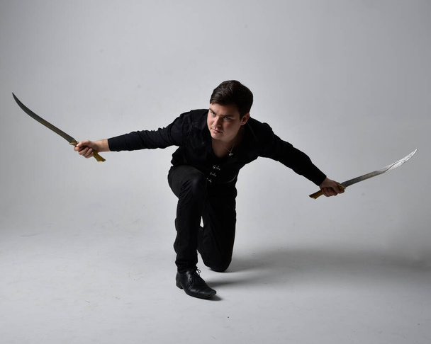 Ganzkörperporträt eines brünetten Mannes in schwarzem Hemd und gotischer Weste mit Schwert. Stehende Action-Pose isoliert vor grauem Studiohintergrund. - Foto, Bild