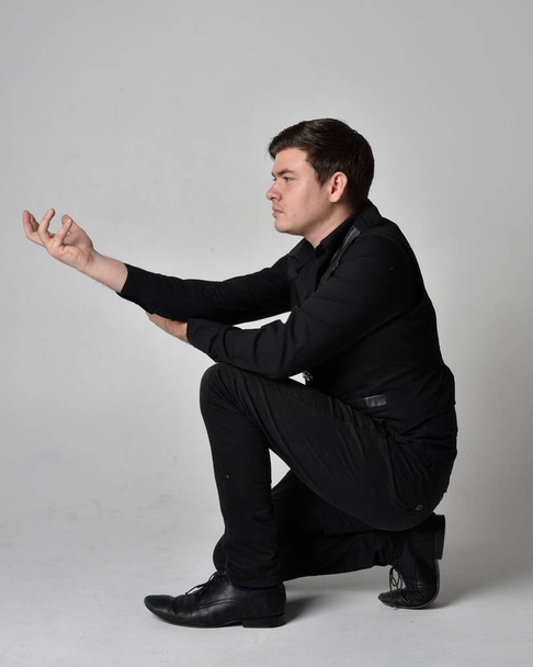 Full length portrait of a  brunette man wearing black shirt and gothic waistcoat.  Seated  pose isolated  against a grey studio background. - Photo, Image
