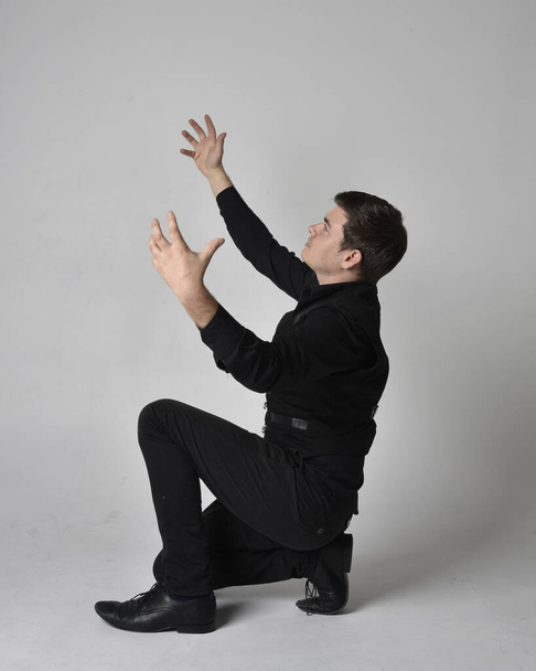 Full length portrait of a  brunette man wearing black shirt and gothic waistcoat.  Seated  pose isolated  against a grey studio background. - Foto, imagen