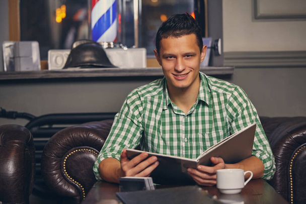 Schöner junger Mann in einem Friseurladen wartet auf den Meister im Wartezimmer, während er Kaffee trinkt - Foto, Bild