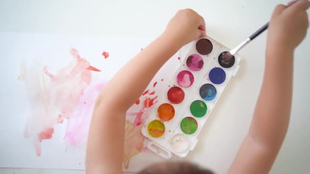 El niño trata de tomar un pincel en la pintura. Un niño pequeño aprende a dibujar en acuarela. Vista superior del proceso de creatividad de los niños. Niño persistente tratando de aprender a dibujar - Metraje, vídeo