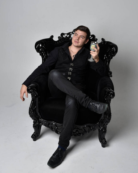 Full length portrait of a  brunette man wearing black shirt and  waistcoat.  Seated  pose in a gothic throne chair isolated  against a grey studio background. - Fotoğraf, Görsel