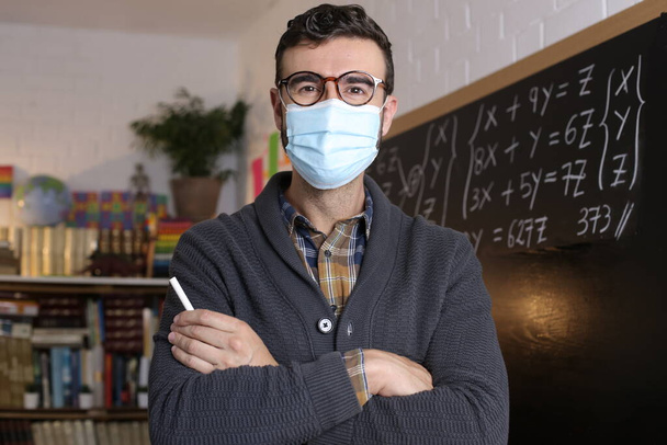 close-up portrait of handsome young teacher with breath mask holding chalk in front of blackboard in classroom - Φωτογραφία, εικόνα