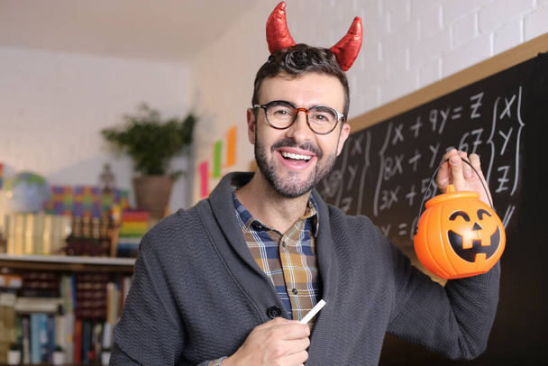 close-up portrait of handsome young teacher with devil horns holding chalk and jack-o-lantern in front of blackboard in classroom - Foto, afbeelding